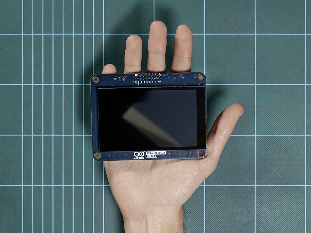 En hand håller en Arduino-pekskärm över en grön rutig bakgrund. Skärmen är avstängd.