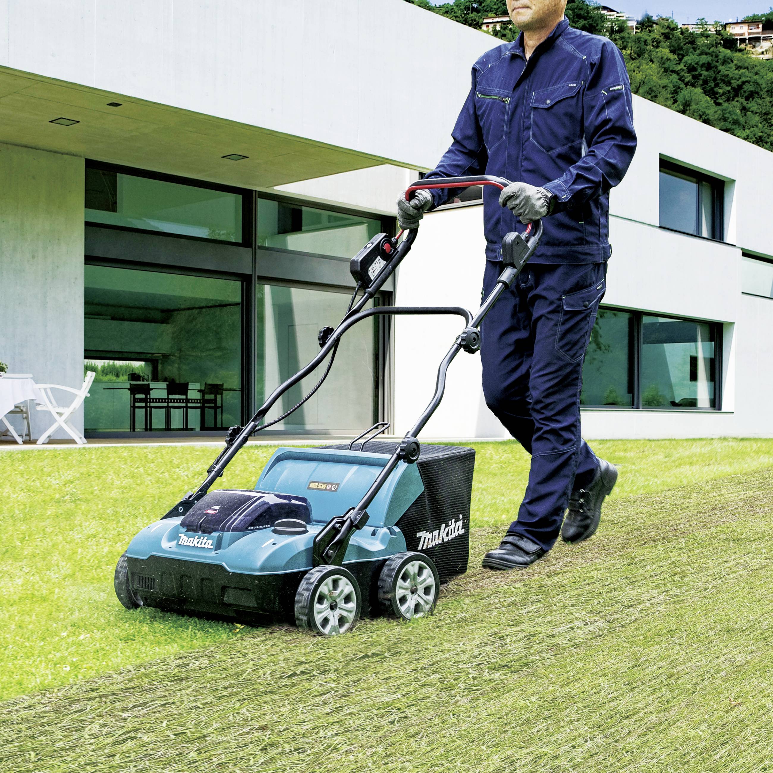 En person använder en vertikalskärare på en gräsmatta framför en modern vit byggnad.