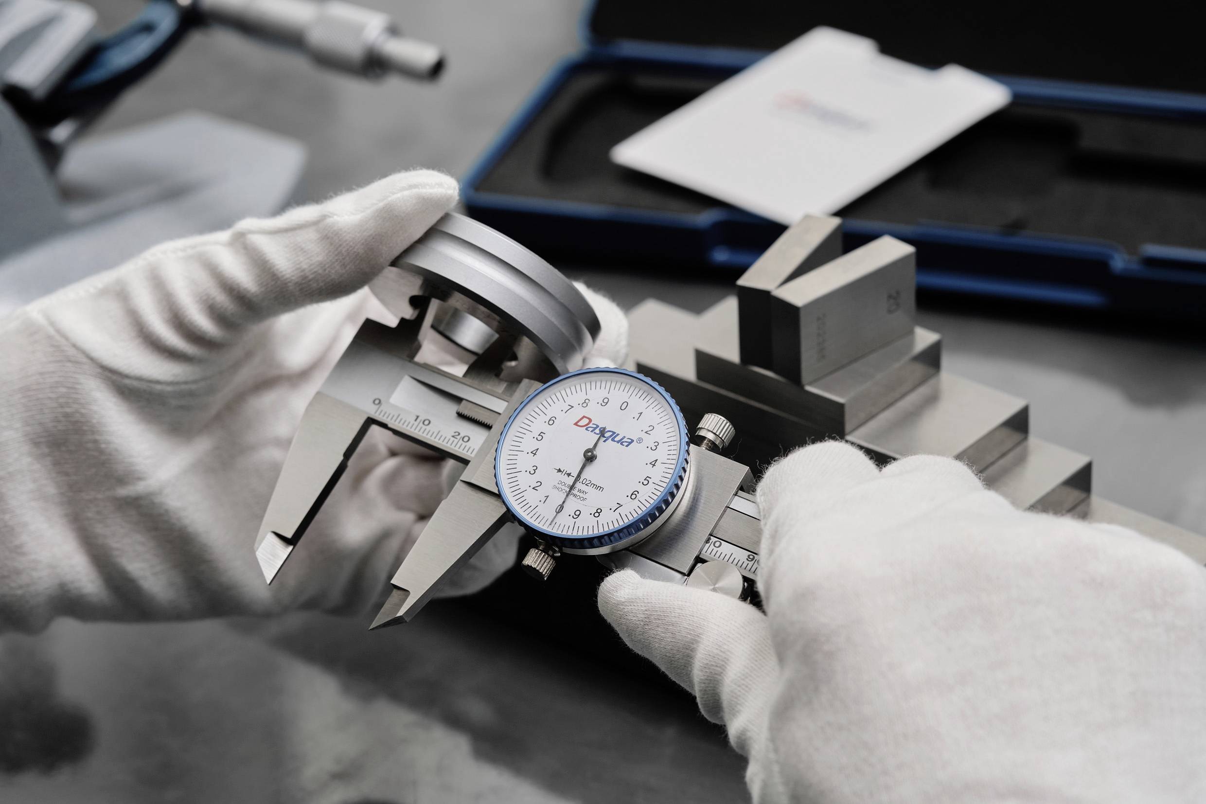 En person mäter ett metallstycke med ett analogt skjutmått. Handskar skyddar mot fingeravtryck. I bakgrunden syns verktyg.