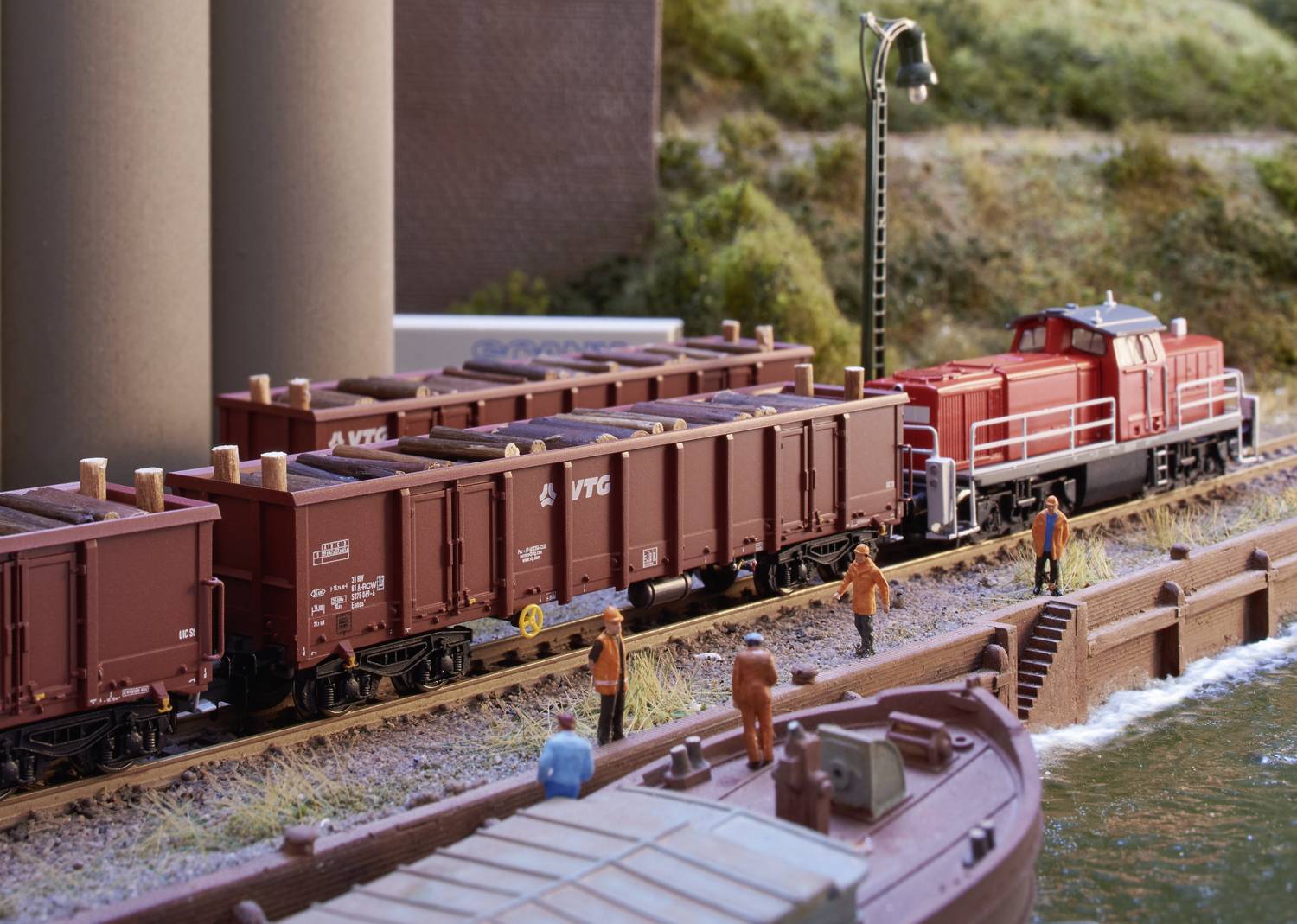 En modelltåg med röda vagnar står på spår bredvid en flod. Fyra miniatyrfigurer syns längs spåren.