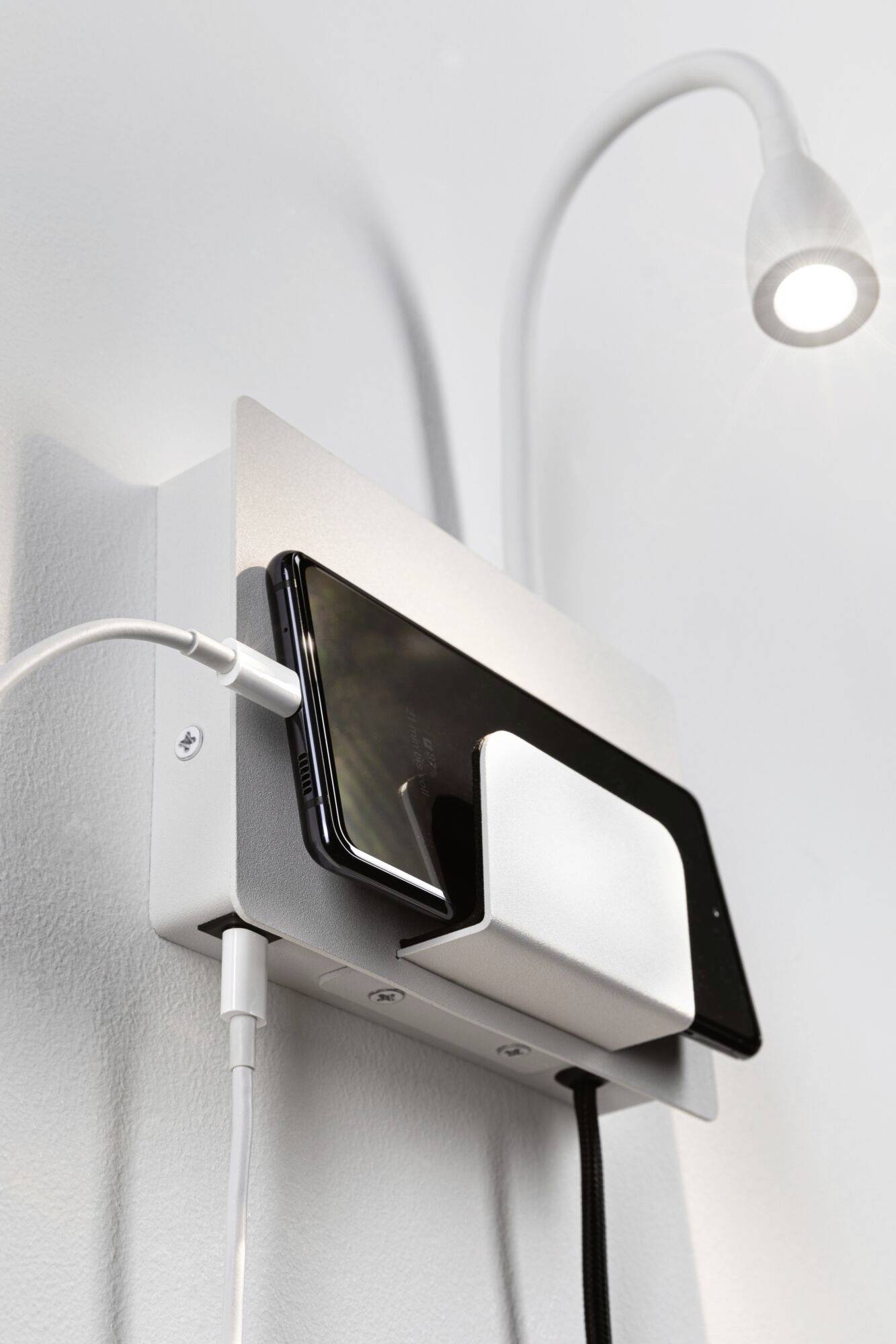 En smartphone laddar trådlöst på en väggmonterad laddare med en integrerad lampa. Två kablar är också anslutna.