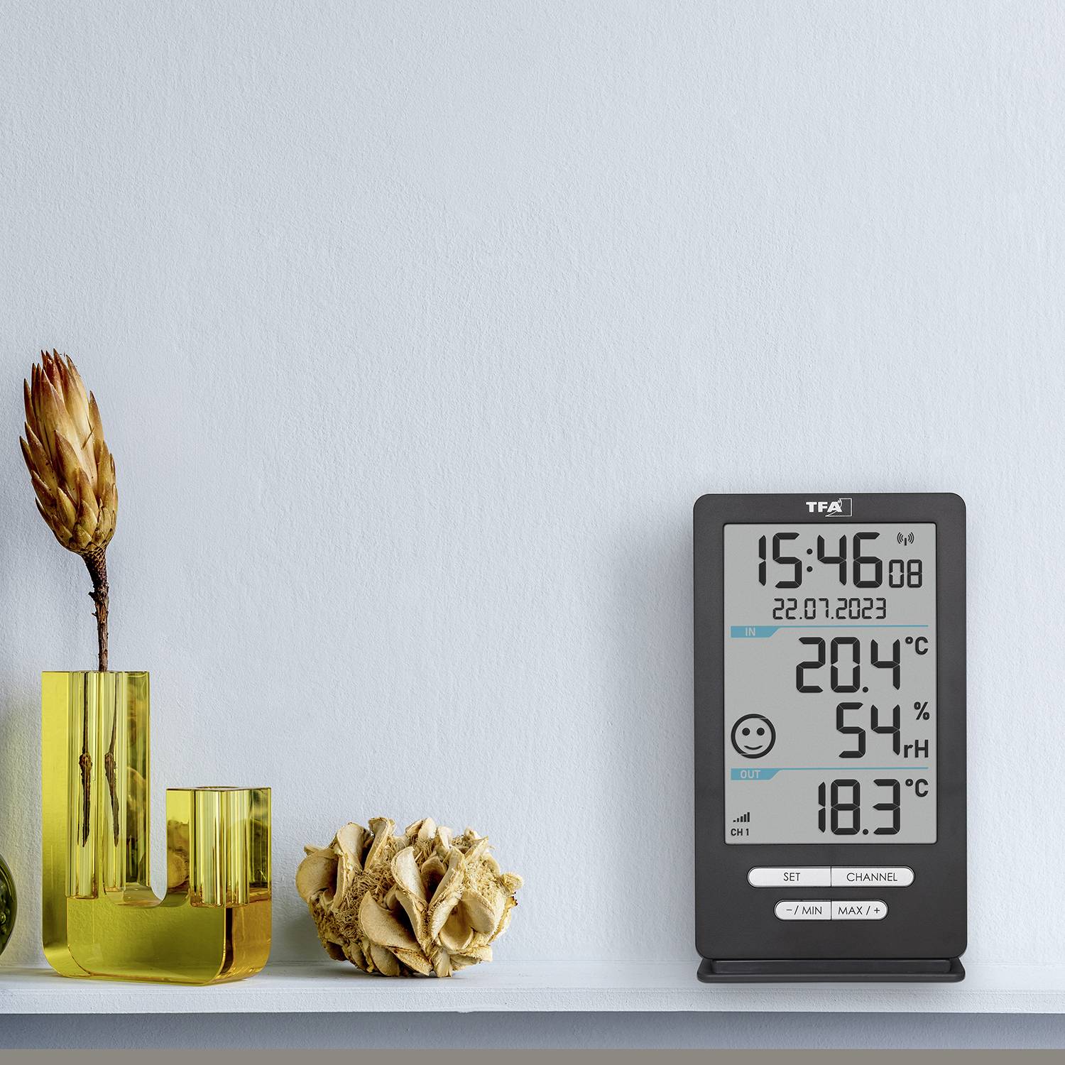 En digital termometer visar temperaturen 20,4°C och luftfuktigheten 54%. Bredvid står en gul vas med en torkad blomma.