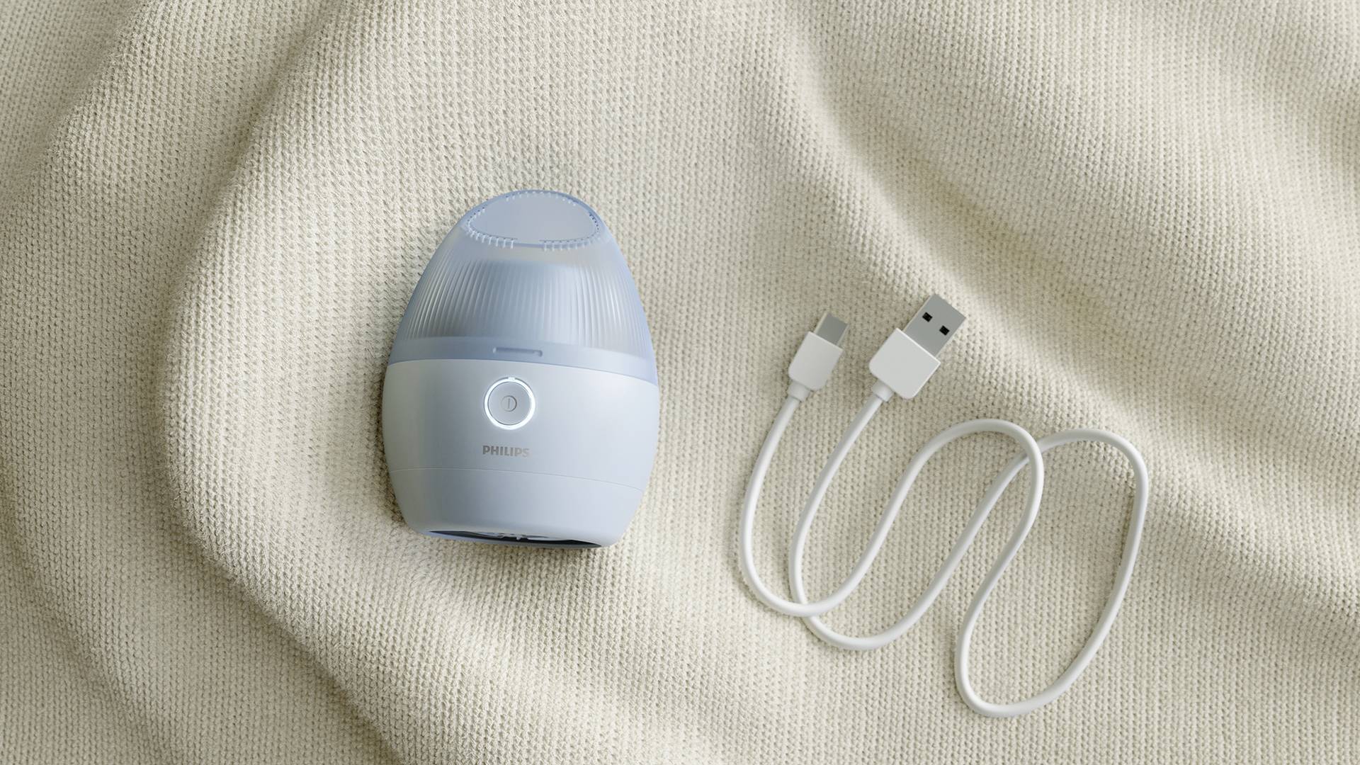 En liten, rund ansiktsmassageapparat med blått hölje och en USB-laddningskabel ligger på en beige tygbakgrund.