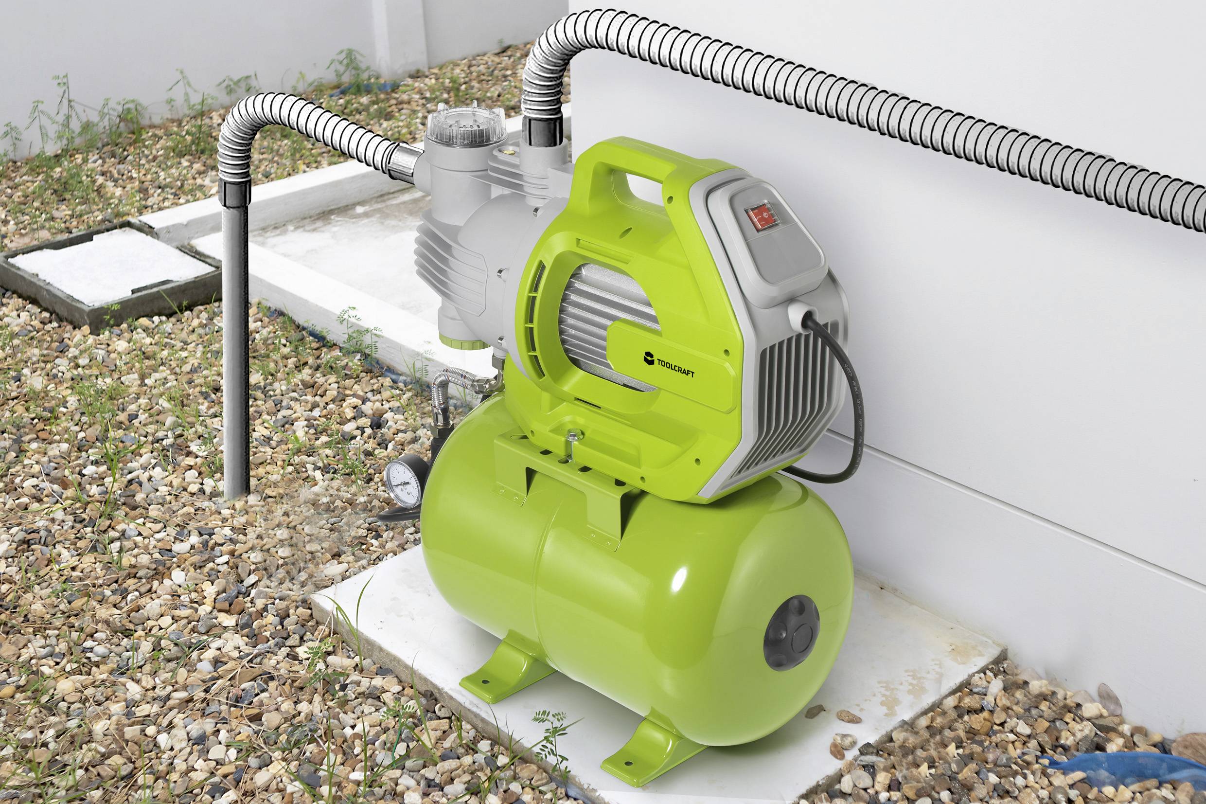 En grön vattenpump med en tank är ansluten till rör. Den står på en grusig mark bredvid en vit vägg.