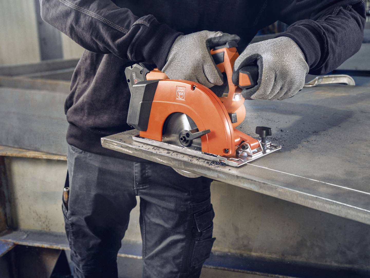 En person i arbetsklädsel och handskar använder en orangefärgad elektrisk handcirkelsåg för att skära ett metallstycke.