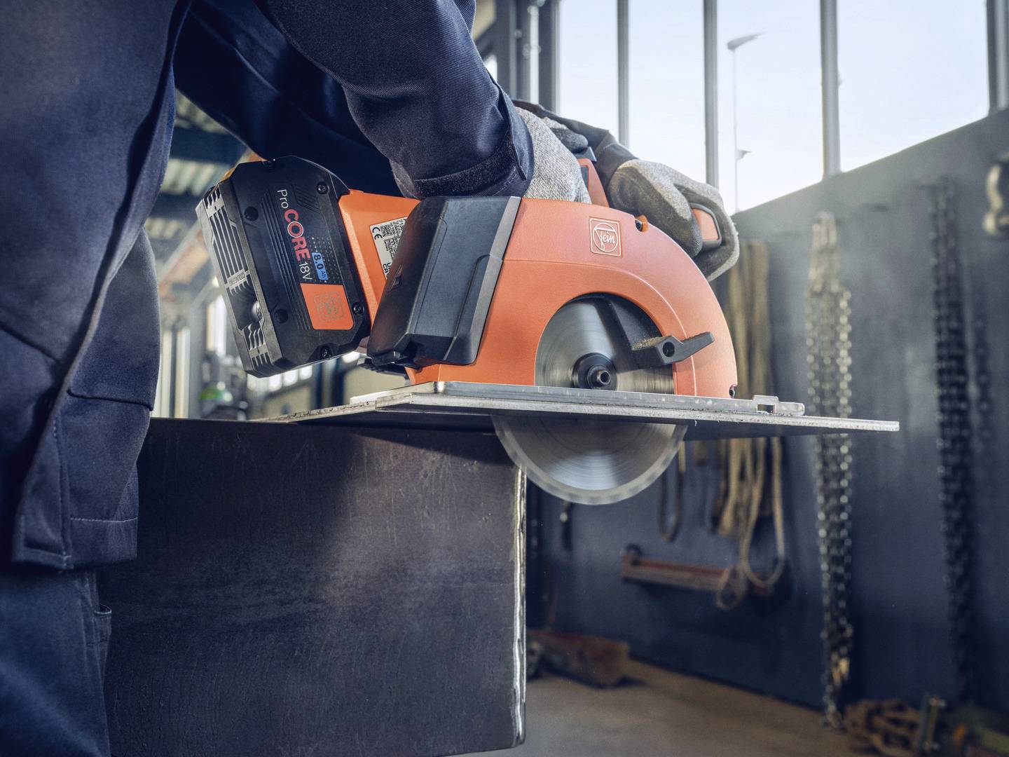 Arbetare klipper metallplatta med en orange batteridriven cirkelsåg i en verkstad, verktyg synliga i bakgrunden.