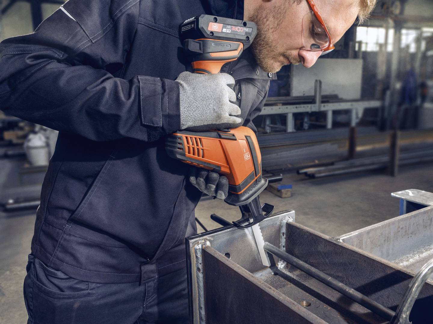 En arbetare i skyddsutrustning sågar metall med en elektrisk såg i en verkstad. I bakgrunden syns industriella maskiner.