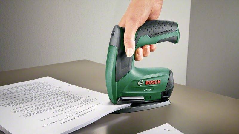 En hand håller en grön Bosch elektrisk häftpistol och fäster papper på ett bord.