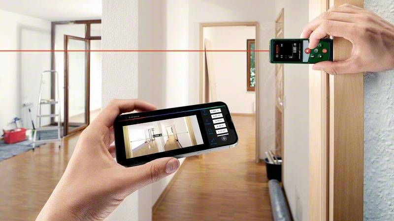 En person mäter längden på ett rum med en lasermätare medan en rumsplan visas på en smartphoneskärm.