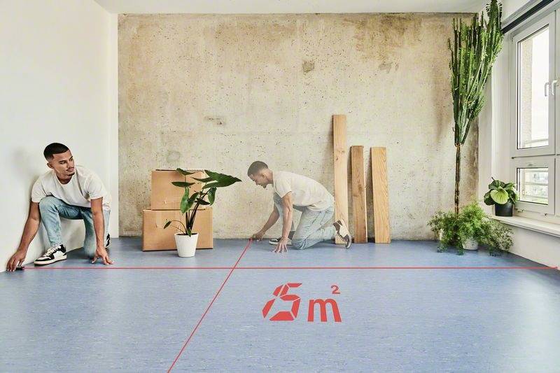 Ett rum med en blå golvyta och ett rött kvadrat markerat '5 m²'. En man mäter ytan. Växter och träbrädor på väggen.