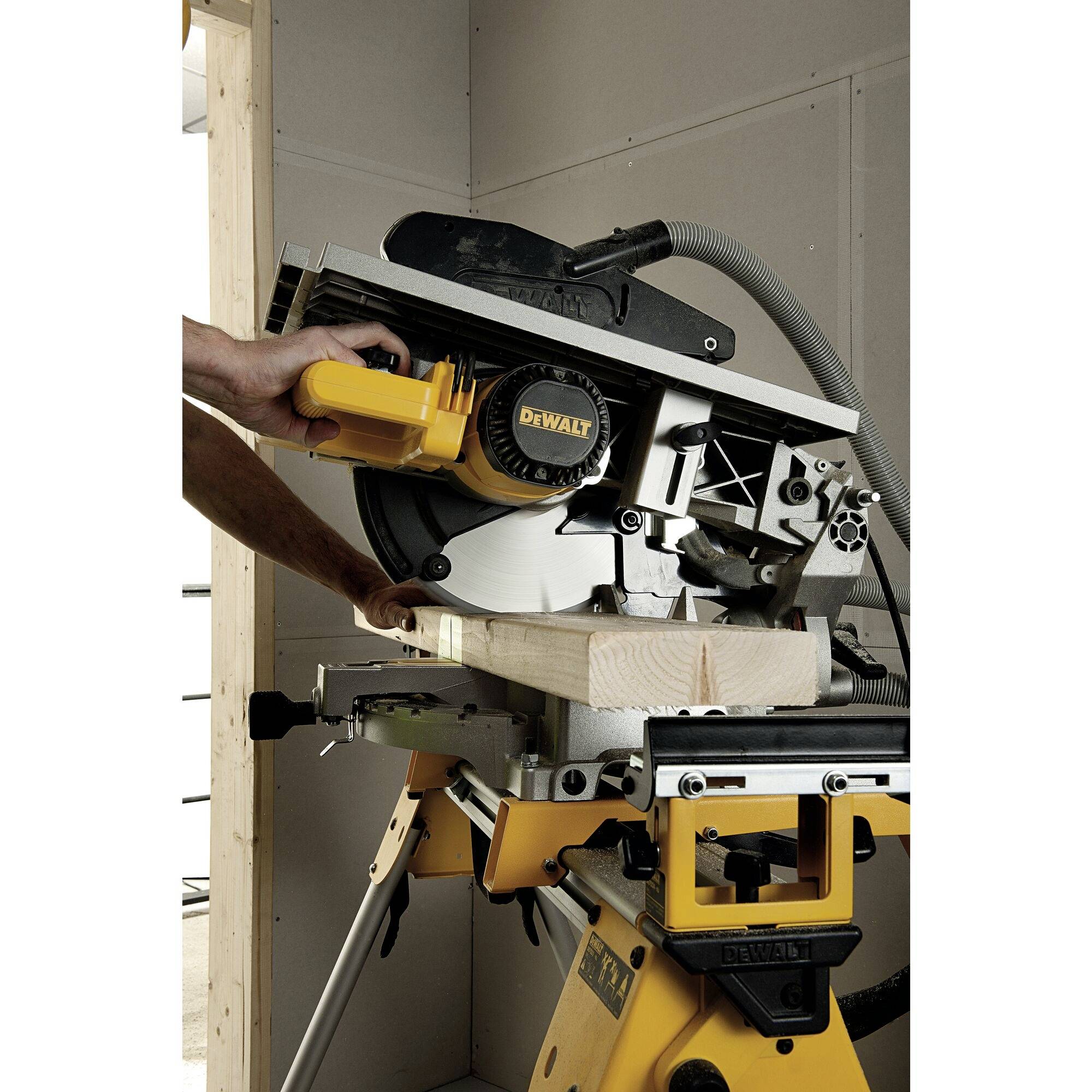 En person använder en kap- och gersåg för att såga ett trästycke. Sågen är gul och har en dammsugeslang.