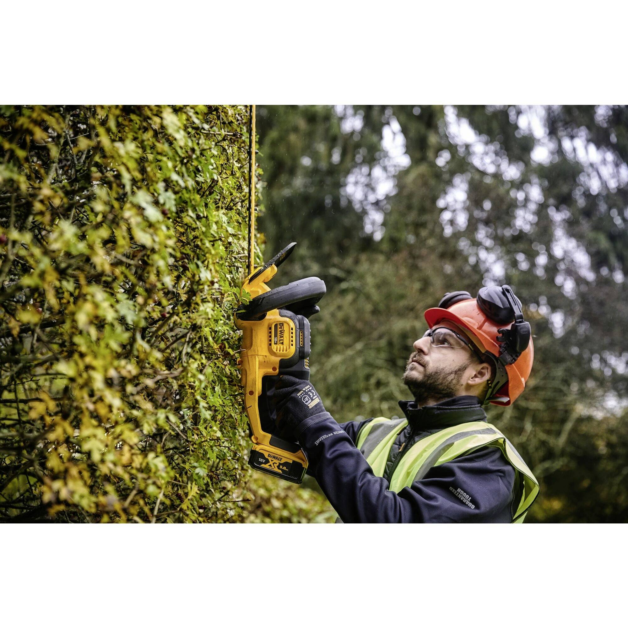 Arbetare med skyddshjälm och handskar klipper en hög häck med elektrisk häcksax; omgiven av träd i bakgrunden.