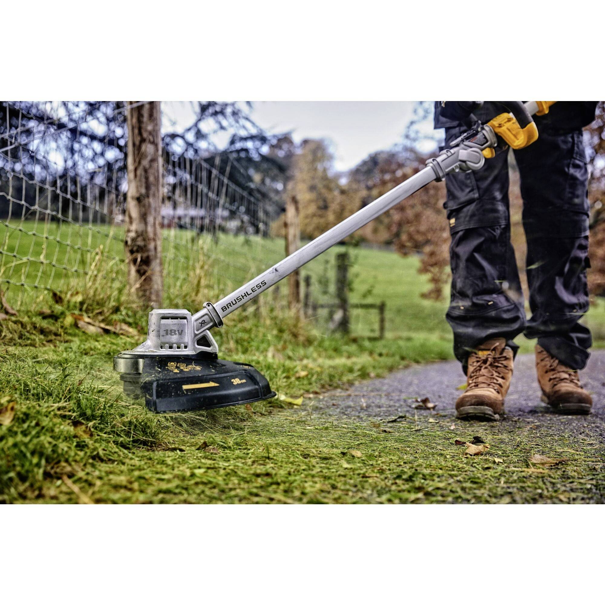 En person klipper gräs med en elektrisk grästrimmer längs en smal väg intill ett staket i en lantlig omgivning.