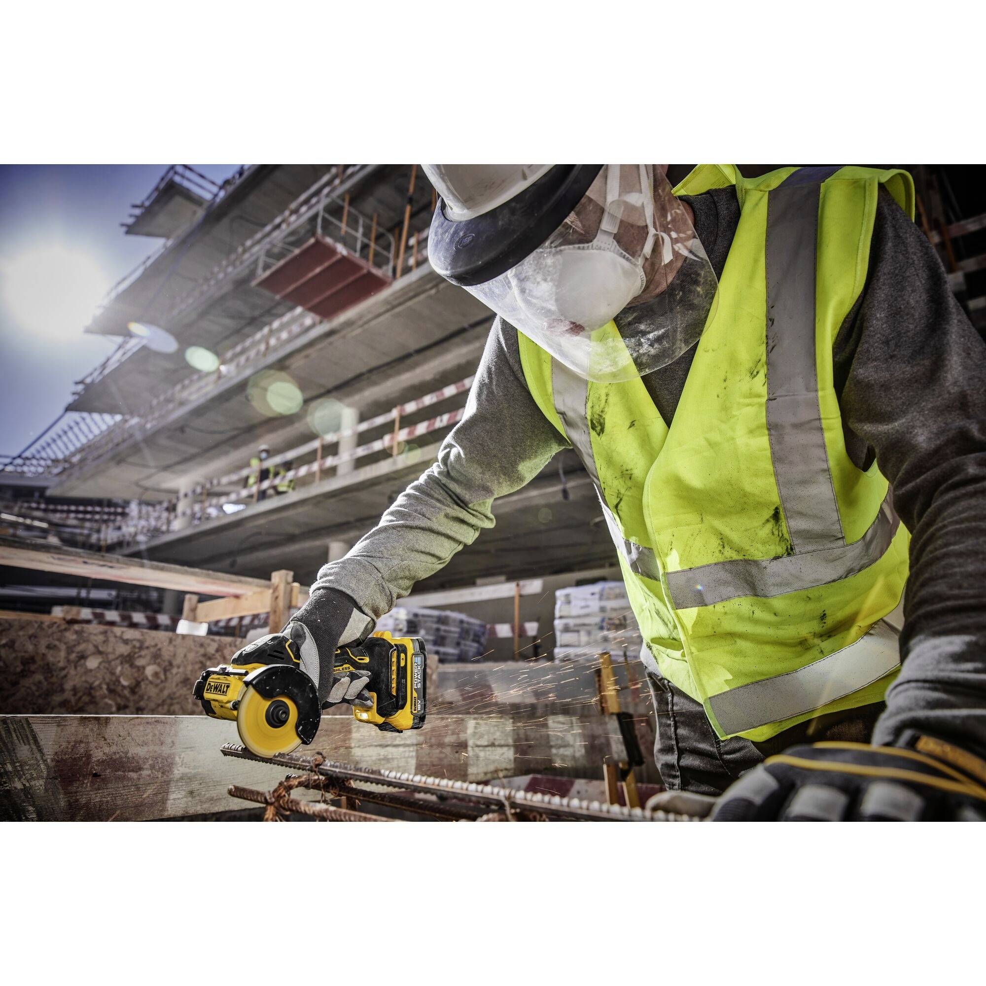 Byggnadsarbetare i skyddsutrustning klipper metallstänger med ett elverktyg på en byggarbetsplats. Bakgrunden visar ett delvis uppfört byggnadsställe.