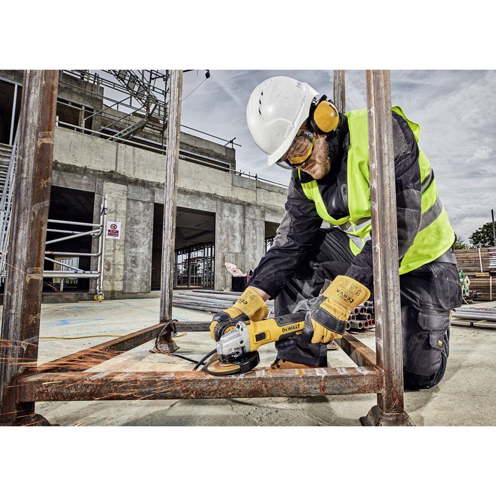 Arbetare med skyddshjälm och skyddskläder slipar metall med vinkelslip på byggarbetsplatsen. Omgivningen består av byggnadsställningar och ett ofärdigt bygge.