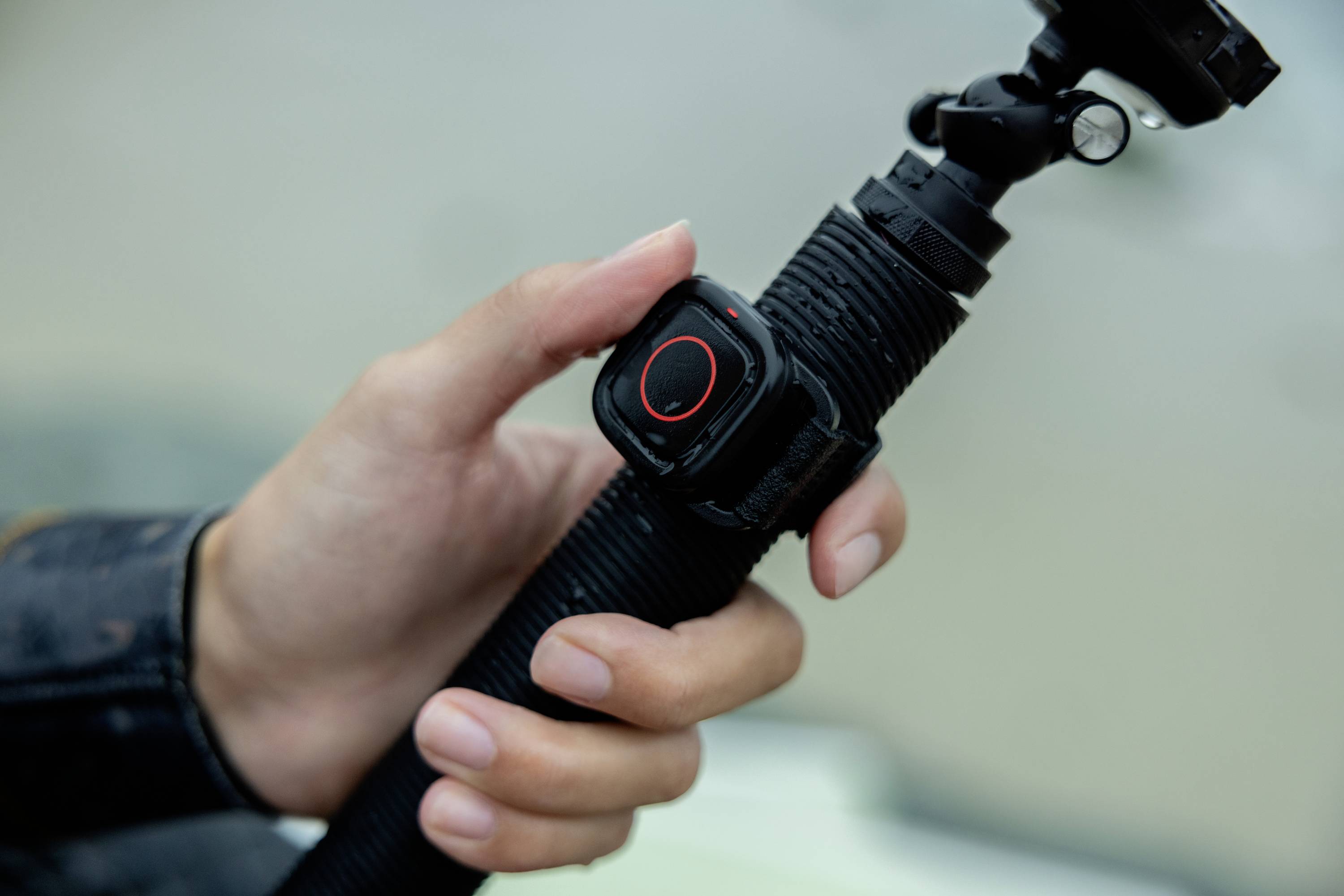 En hand håller ett svart selfie-stick med en liten, fyrkantig enhet längst upp som visar en påslagningsknapp med röd kant.
