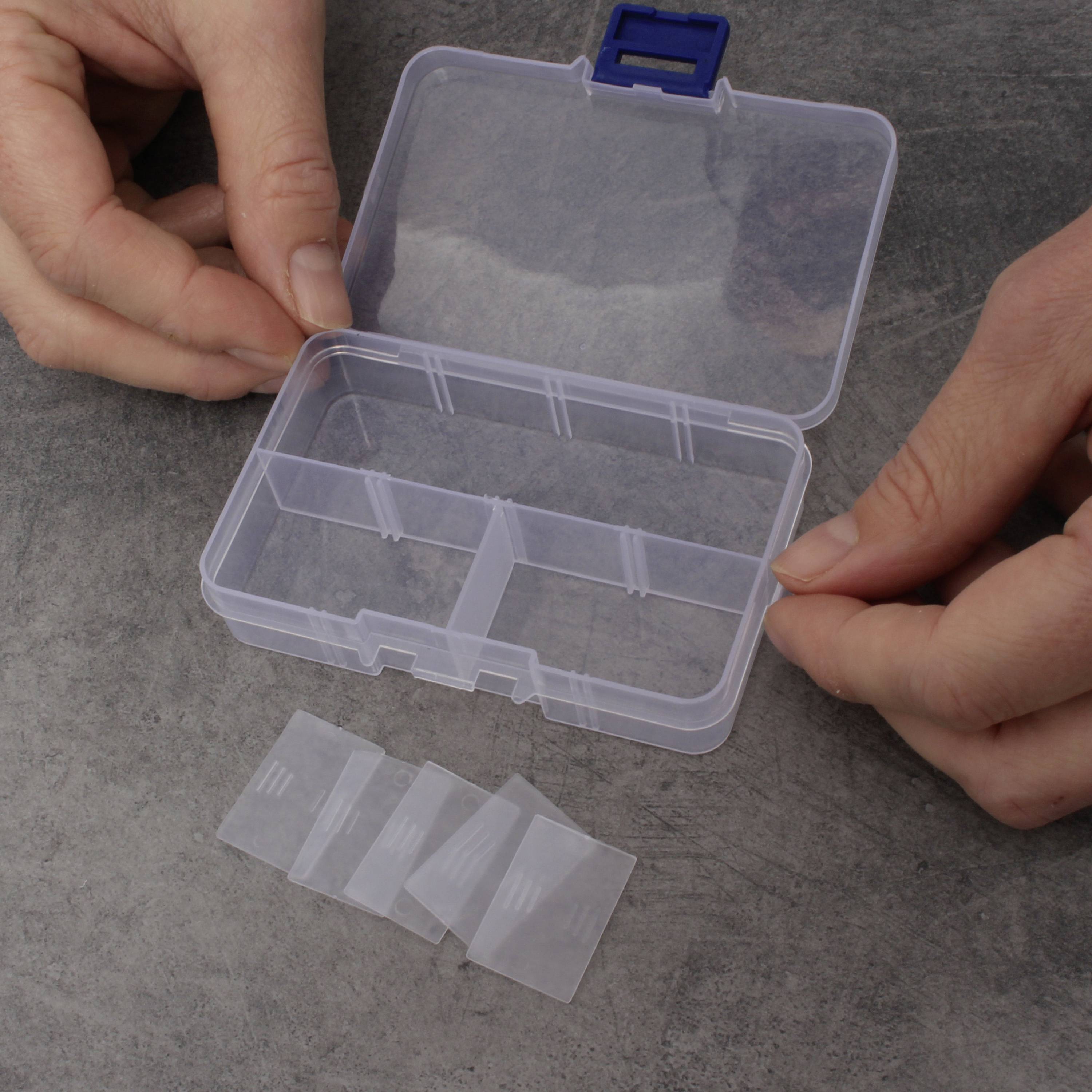 Två händer öppnar en liten genomskinlig plastbehållare med flera fack. Fyra skiljeväggar ligger bredvid på ett bord.