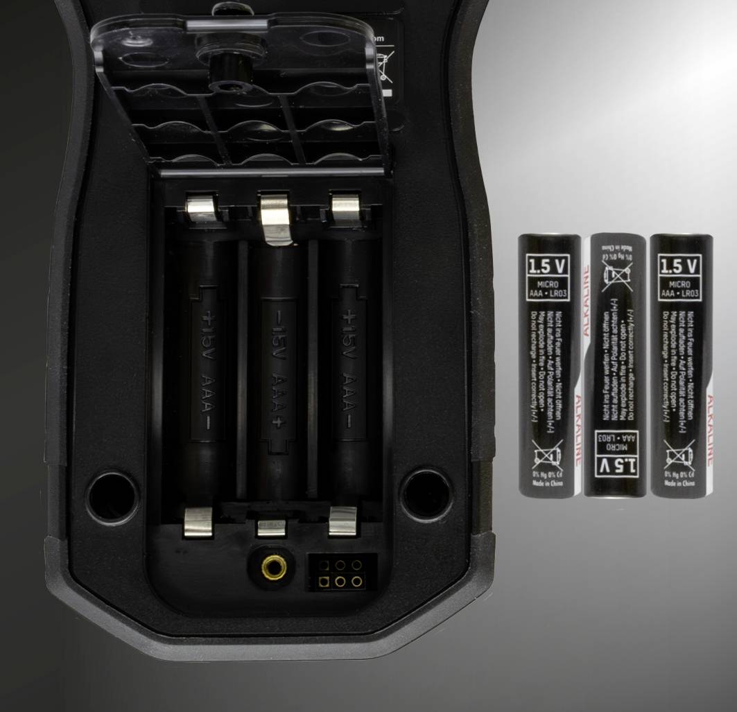 Ett öppet batterifack på en enhet som visar tre tomma AA-batterifack; bredvid finns tre AA-batterier på 1,5 V.