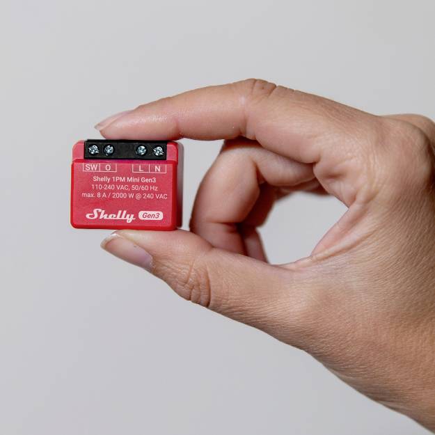 En hand håller en liten röd enhet som heter 'Shelly Gen3' med elektriska anslutningsklämmor och tekniska specifikationer på framsidan.