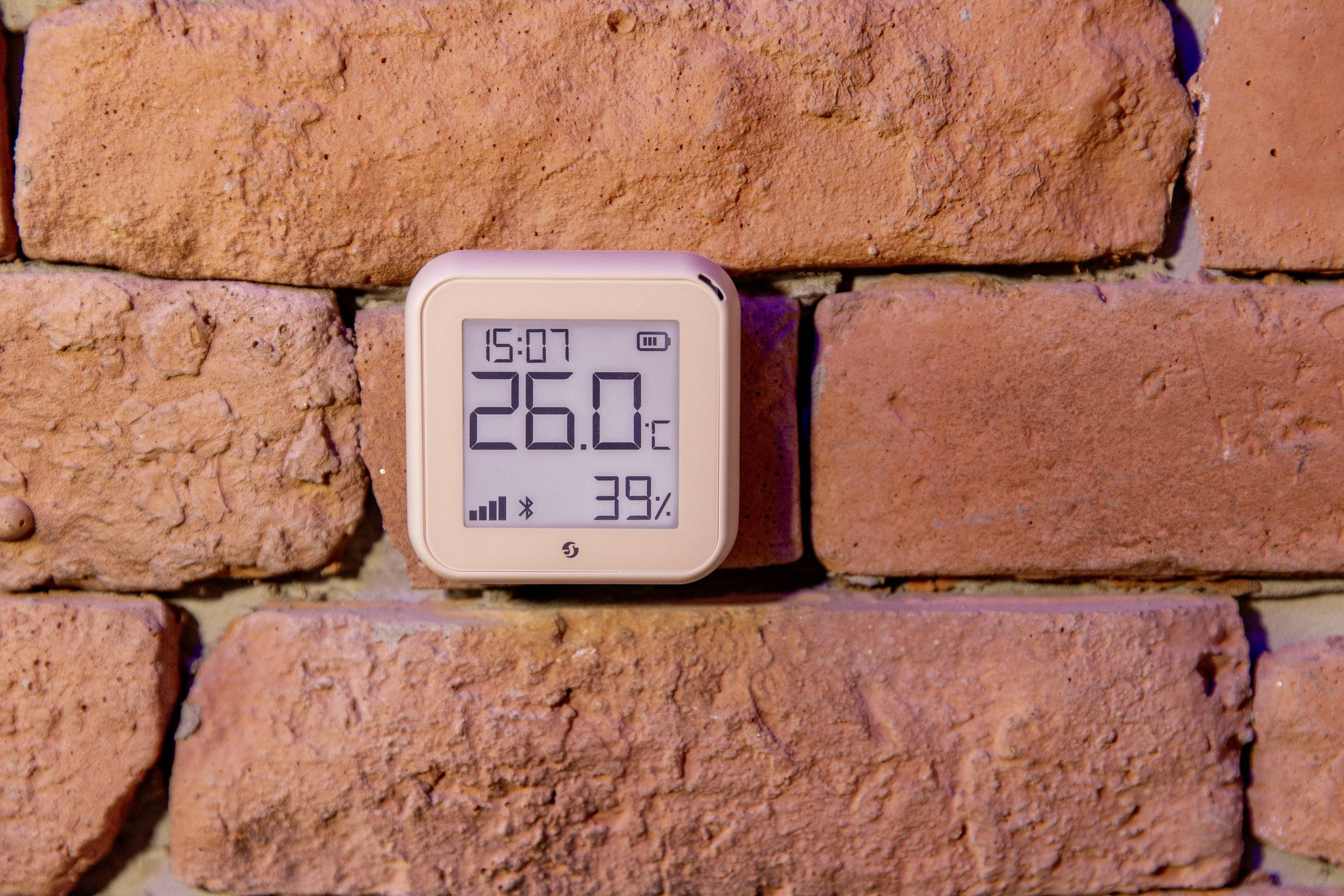 Smart-termostat på en tegelmur visar 15:07, 26,0°C och 39% luftfuktighet.