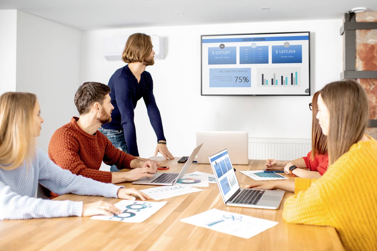 En grupp på fem personer sitter runt ett bord och betraktar en skärm med finansdiagram och data i ett mötesrum.