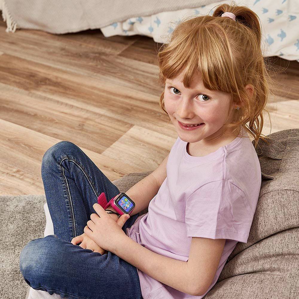 Ett barn sitter på golvet, bär en lila t-shirt och blå jeans. Det tittar leende in i kameran och håller en smartwatch i handen.