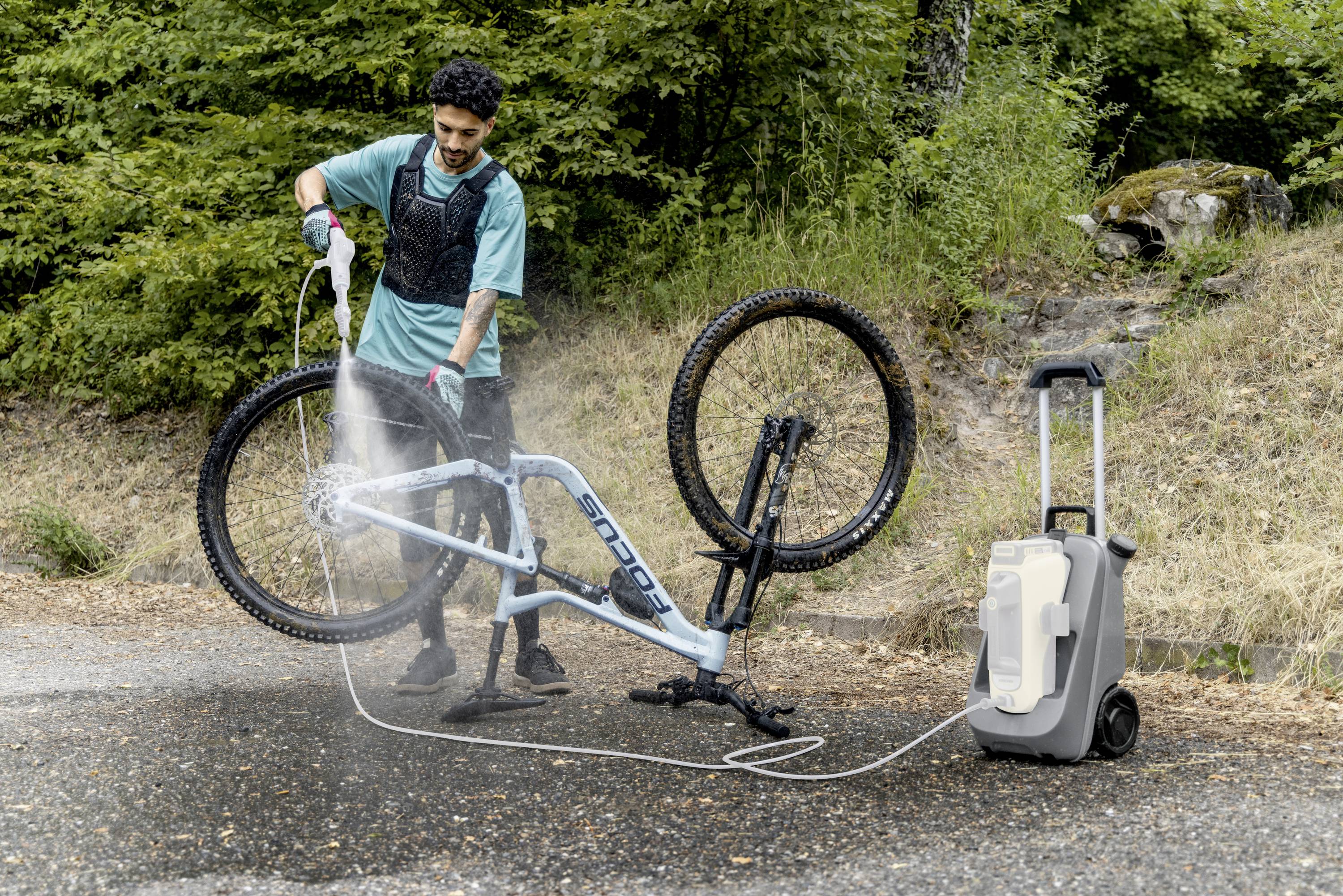 En person rengör en upp-och-nervält cykel med en högtryckstvätt utomhus. I bakgrunden syns träd.