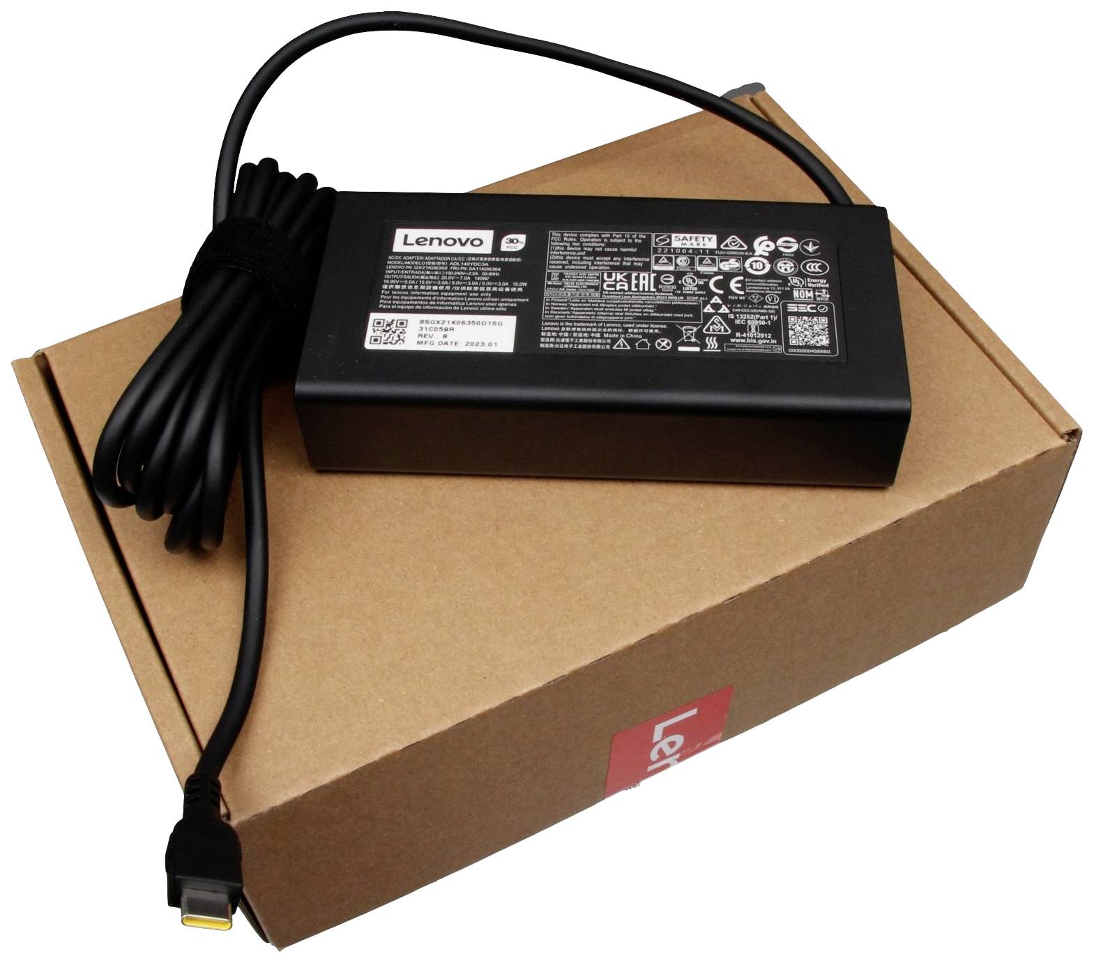 Ett svart Lenovo-nätadapter ligger på en brun kartongförpackning. På förpackningen syns tekniska detaljer och logotyper.