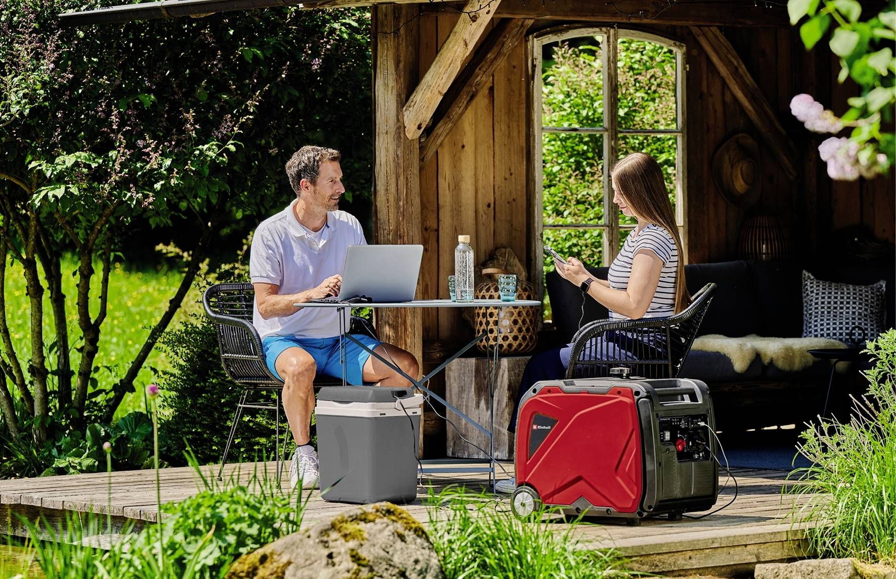 En kvinna och en man sitter på en terrass i trädgården och arbetar på laptops, en röd generator står bredvid.
