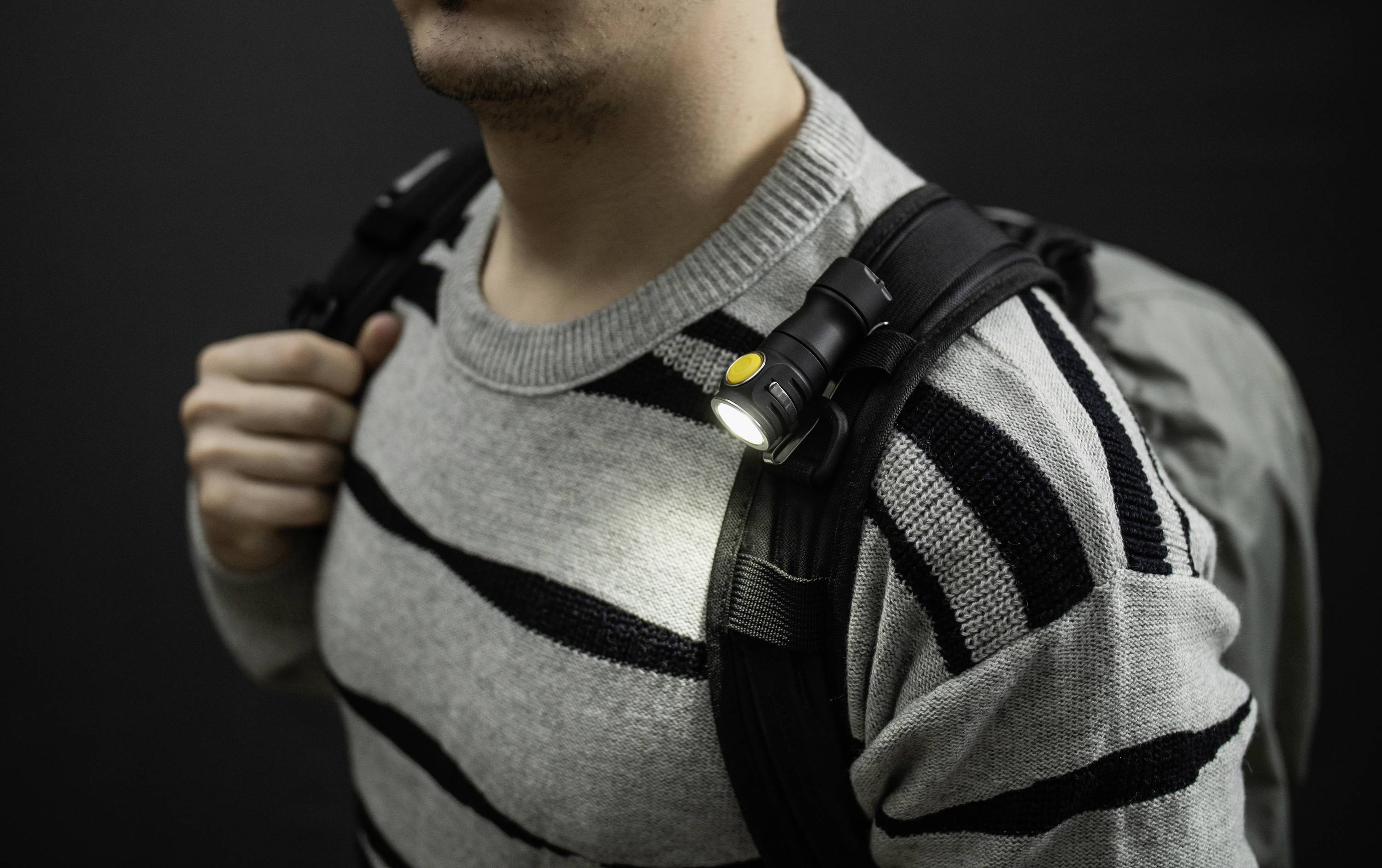 En person bär en grå ryggsäck med ett lysande litet ljus fäst vid axeln mot en mörk bakgrund.