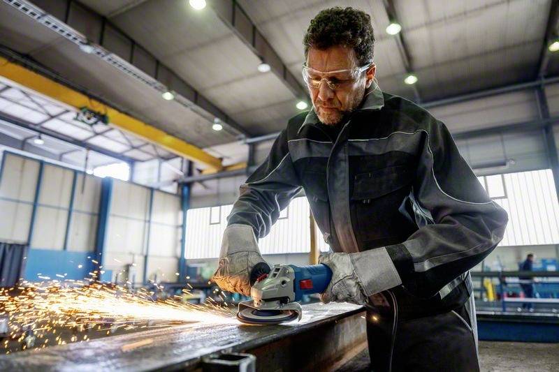 En arbetare sliper metall med en vinkelslip, gnistor flyger. Han bär skyddsglasögon och handskar. Fabriksmiljö i bakgrunden.