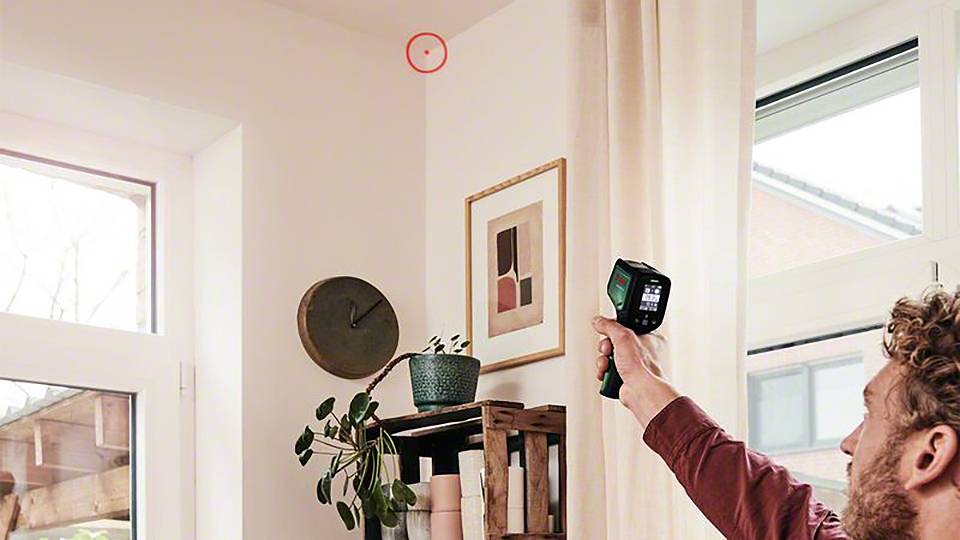En person skannar en väggörn med en värmekamera i ett bostadsutrymme. En röd cirkel markerar en punkt i taket.