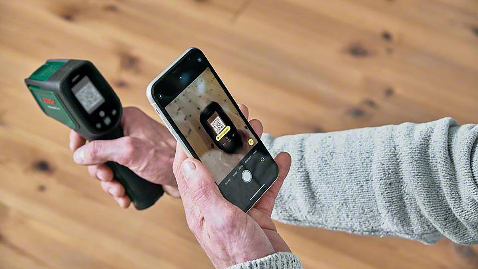En person håller ett infraröd-termometer och en smartphone som fotograferar termometerns skärm. Bakgrunden är ett trägolv.