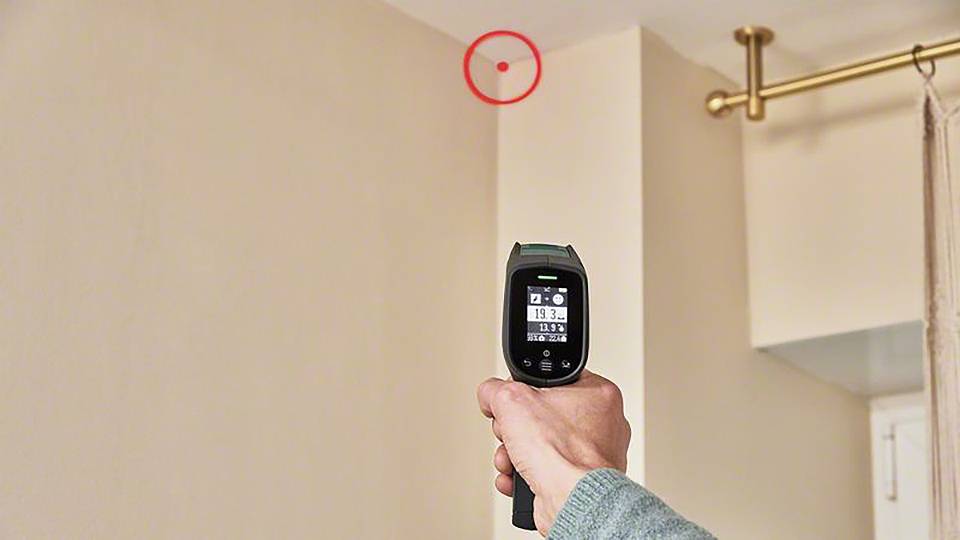 En person siktar med en infraröd termometer mot ett rumstak. En röd cirkel markerar en punkt på taket.