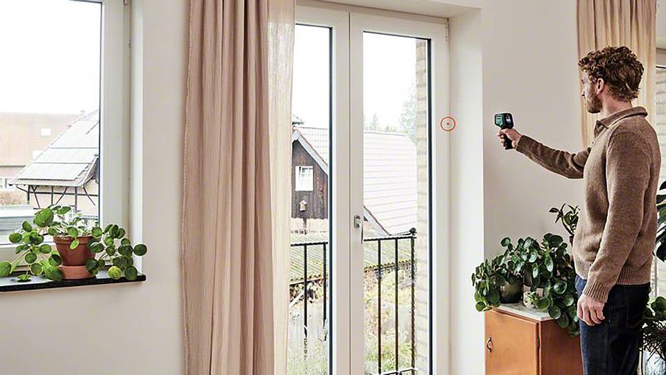 En man mäter isoleringen på ett stort fönster med en apparat. Växter står på fönsterbrädan och ett skåp.