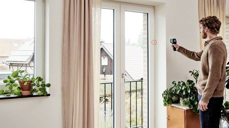 En man mäter utomhustemperaturen genom en stängd terrassdörr med hjälp av en mätenhet. I bakgrunden syns solpaneler.