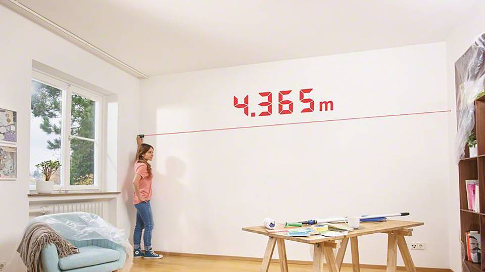 En kvinna mäter bredden på en vit vägg med ett digitalt måttband; displayen visar '4,365 m'.