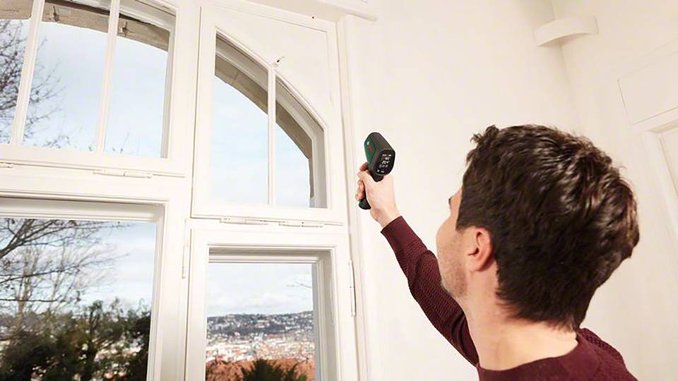 En man mäter temperaturen vid ett fönster i ett ljust rum. Genom fönstret syns ett stadslandskap.