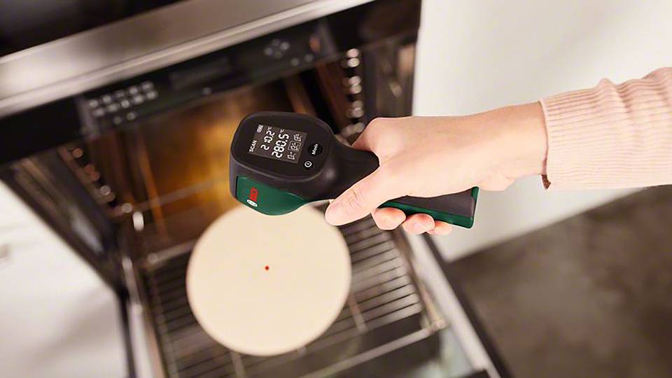 En person håller ett infraröd-termometer över en öppen ugnslucka för att mäta temperaturen på en pizzasten.
