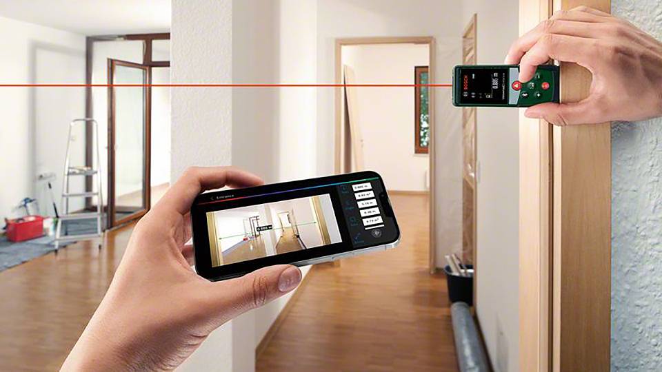 Två händer håller ett digitalt lasermätinstrument och en smartphone med en app som visar rumslig mätning. I bakgrunden ett rum.