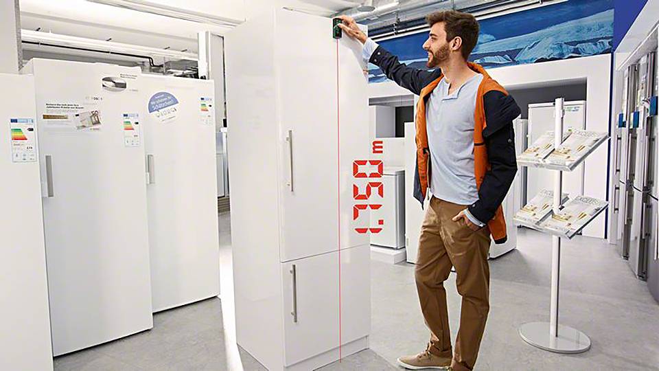 En man mäter höjden på ett kylskåp med ett lasermätverktyg i en hushållsapparatbutik.