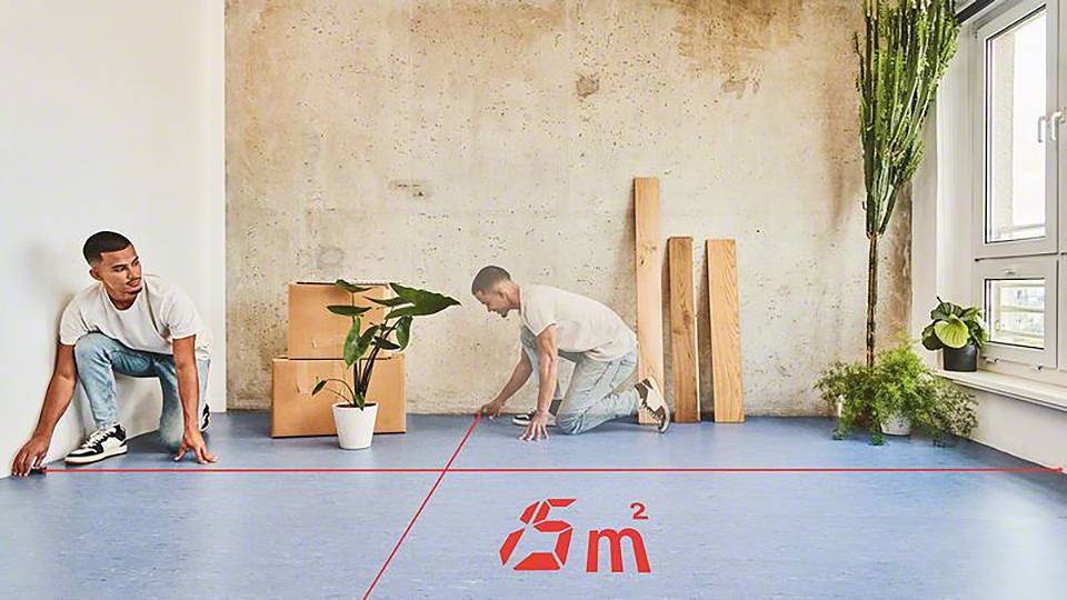 Två män med en växt och träbitar markerar ett område på ett golv. Området är märkt med '5 m²'.