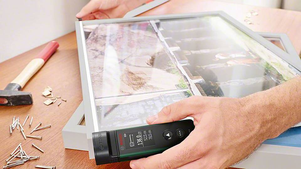 En person riktar in en ram med en digital vinkelmätare på ett bord. En hammare och spikar ligger bredvid.