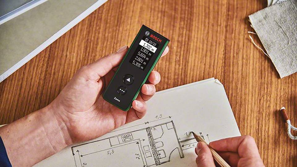 En hantverkare mäter distansen med ett Bosch-lasermätinstrument på ett bord med byggnadsritningar. Displayen visar '1,823 m'.