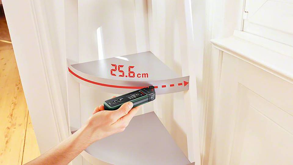 En hand håller en digital mätare i ett rum och mäter ett avstånd på 25,6 cm vid en vit vägg.