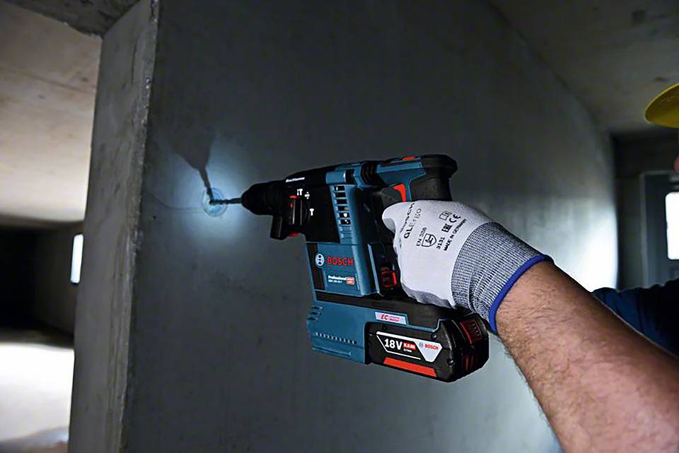 En person använder en blå Bosch batteridriven slagborrmaskin mot en betongvägg inomhus. Personen har en skyddshandske på sig.