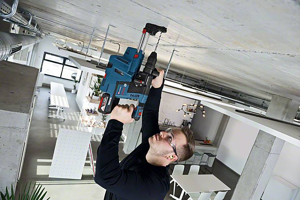 En person borrar i ett betongtak med en stor, blå borr i en industriell miljö. I bakgrunden syns moderna möbler.