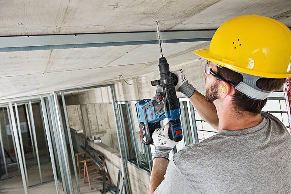 En byggnadsarbetare med skyddshjälm och hörselskydd borrar i ett tak med en elektrisk borrmaskin.