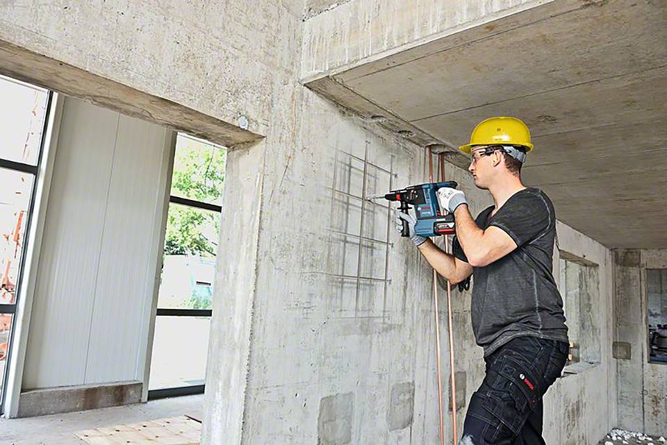 En byggnadsarbetare med gul hjälm borrar i en betongvägg med en elektrisk borr på en byggarbetsplats.