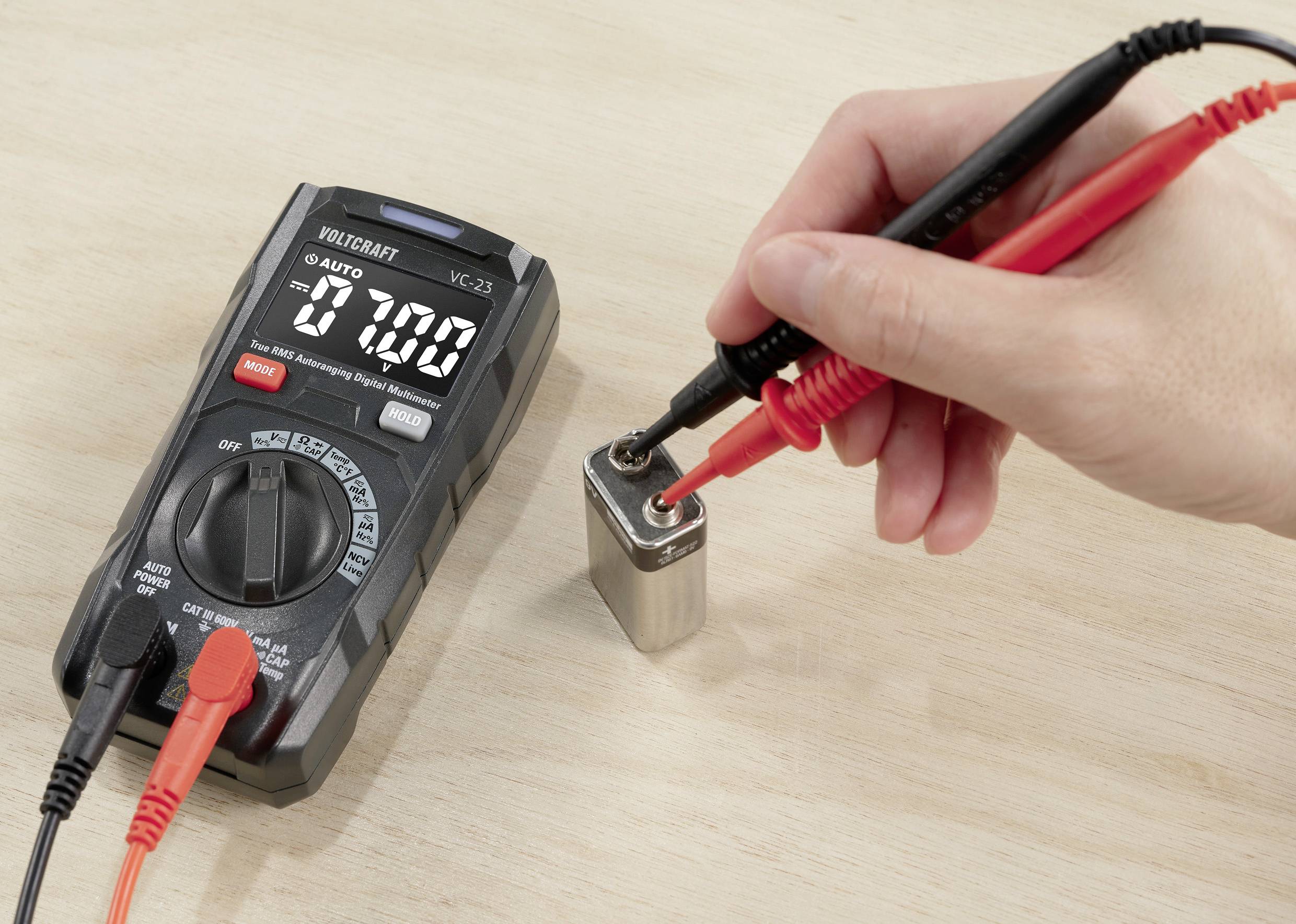 Hand med multimeterns mätspetsar mäter spänningen på ett 9-volts batteri. Multimetern visar 7,00 volt. Träyta i bakgrunden.